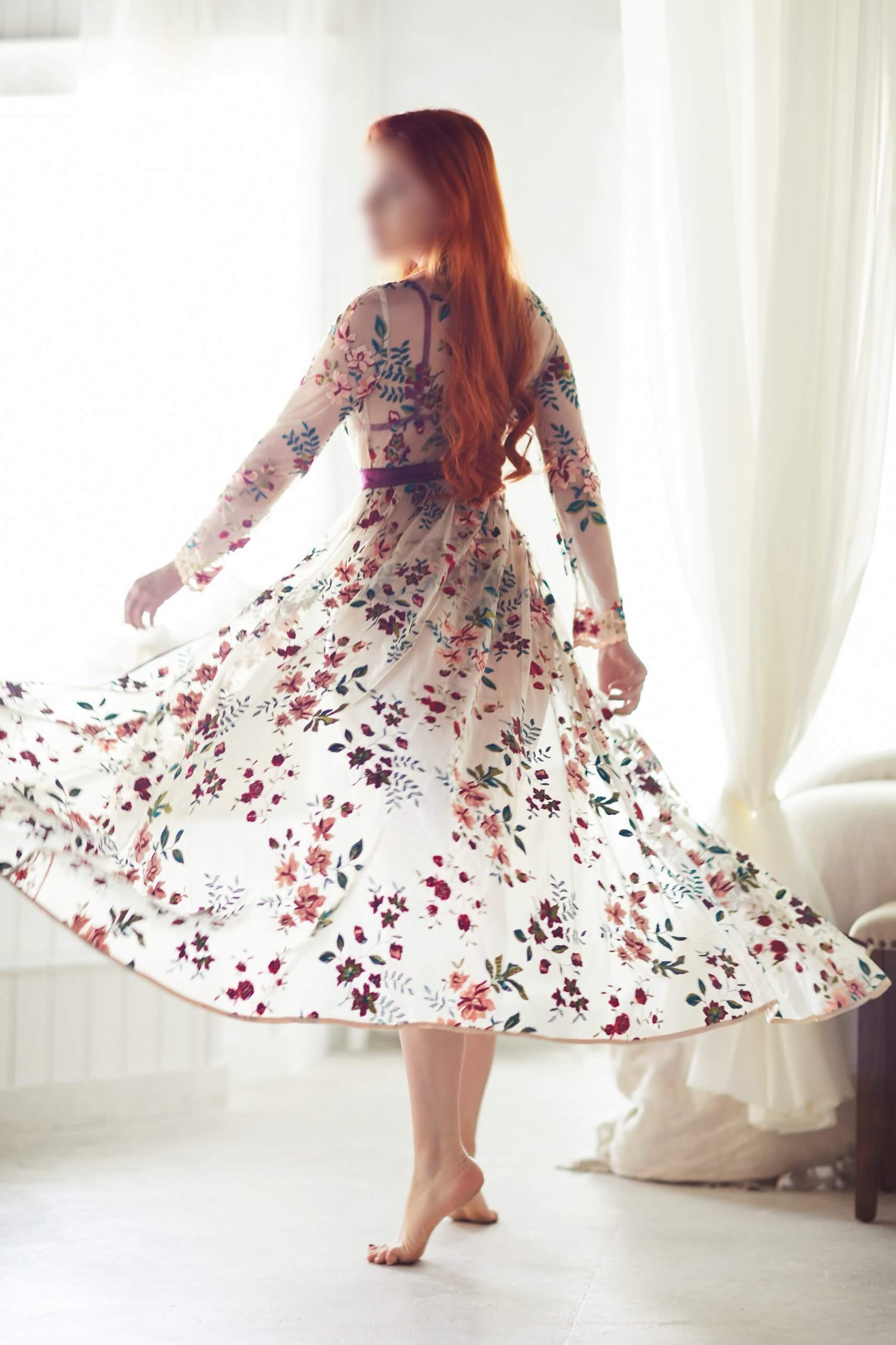 Redhead escort Josilee Adams spins in a sheer floral robe, arms on either side of her slender body.