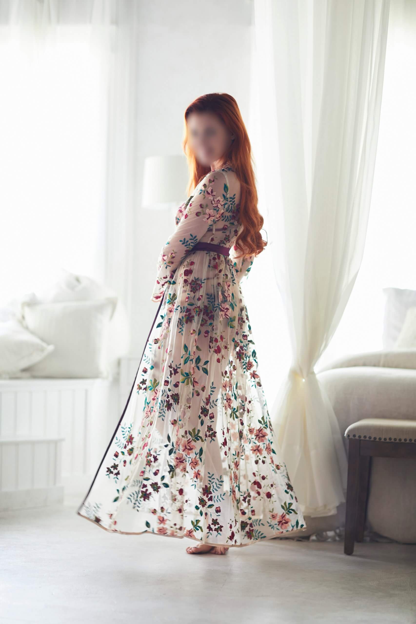 Bathed in natural light, Josilee Adams exudes soft romance in a sheer floral gown. Her flowing red hair complements the delicately embroidered dress as she stands barefoot, framed by gauzy white curtains in a dreamy bedroom.