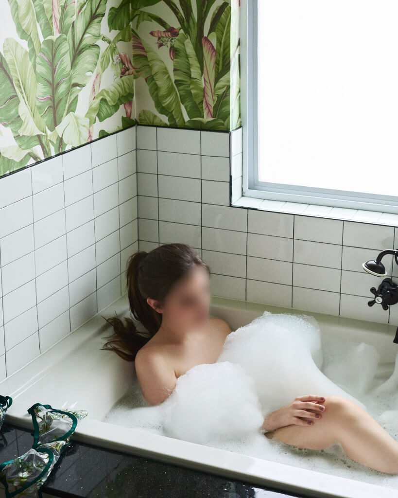 Josilee Adams relaxes in a deep bubble bath, surrounded by white tile and banana leaf wallpaper, her lingerie draped over the edge of the tub in a warm, steamy glow.
