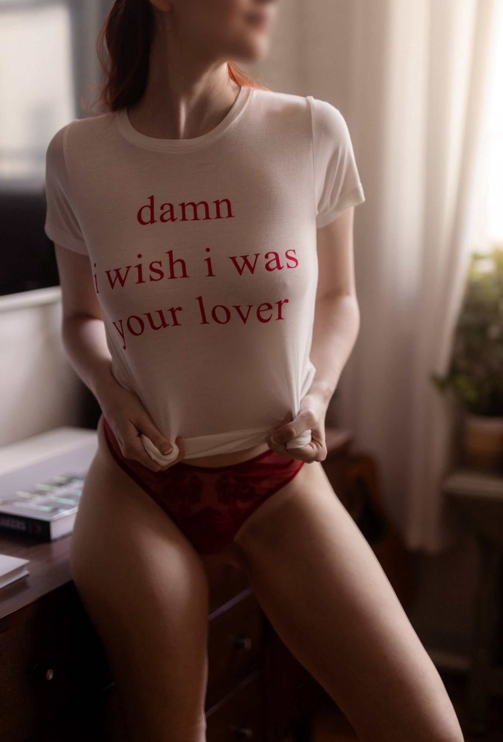 Redhead escort Josilee Adams playfully tugs at her white shirt, bold red text reading