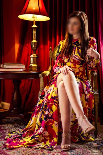 Josilee Adams sits regally on an ornate chair in a richly colored room, wearing a long floral dress with crossed legs and soft lighting from a table lamp.